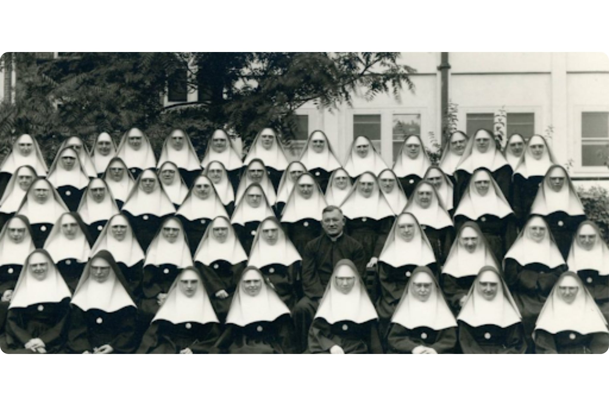 A historic, wide-angle group photograph showing rows of Sisters of The Sorrowful Mother, seated outdoors with a priest in the center.