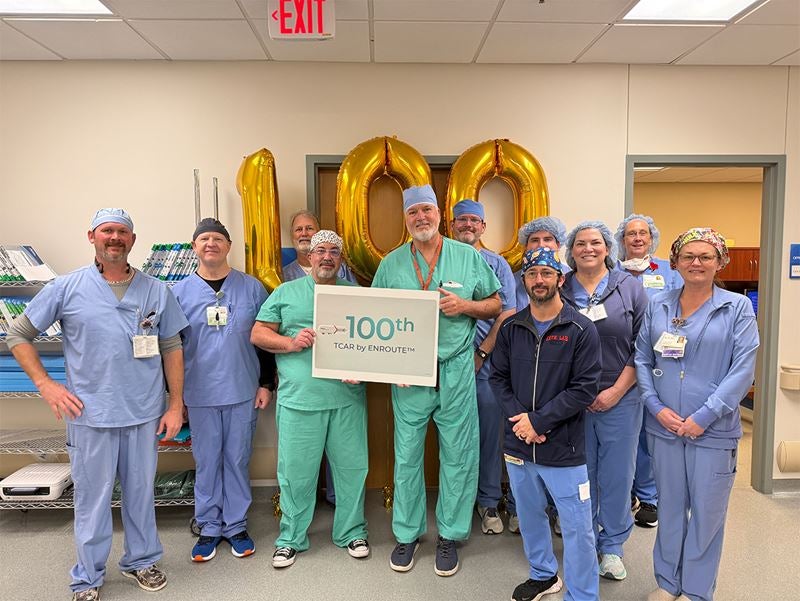 Group of surgical staff in scrubs stand in a hospital room in front of gold “100” balloons, holding a sign marking the 100th TCAR stroke-prevention procedure performed by Dr. Geoffrey Risley, celebrating the milestone with the care team.