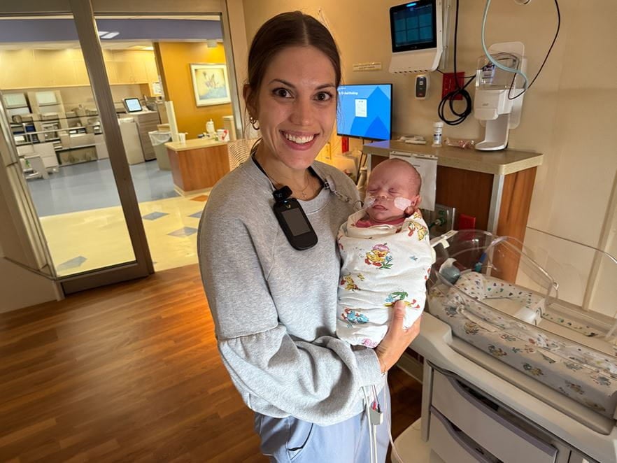Hana Scott, RN, a neonatal ICU nurse at Ascension Columbia St. Mary’s Milwaukee Campus, smiles while holding a swaddled newborn in a hospital patient room with medical equipment nearby.