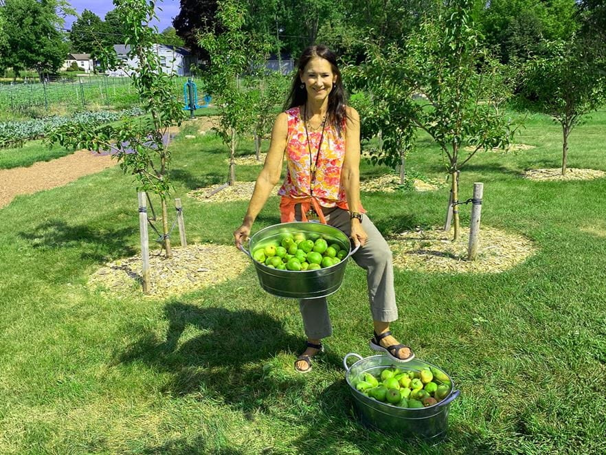 Jenny Ovide, RN, of Community Medicine at Ascension All Saints Family Health Center, stands in a garden holding a tub of freshly picked green apples, with another basket of apples at her feet and fruit trees in the background.