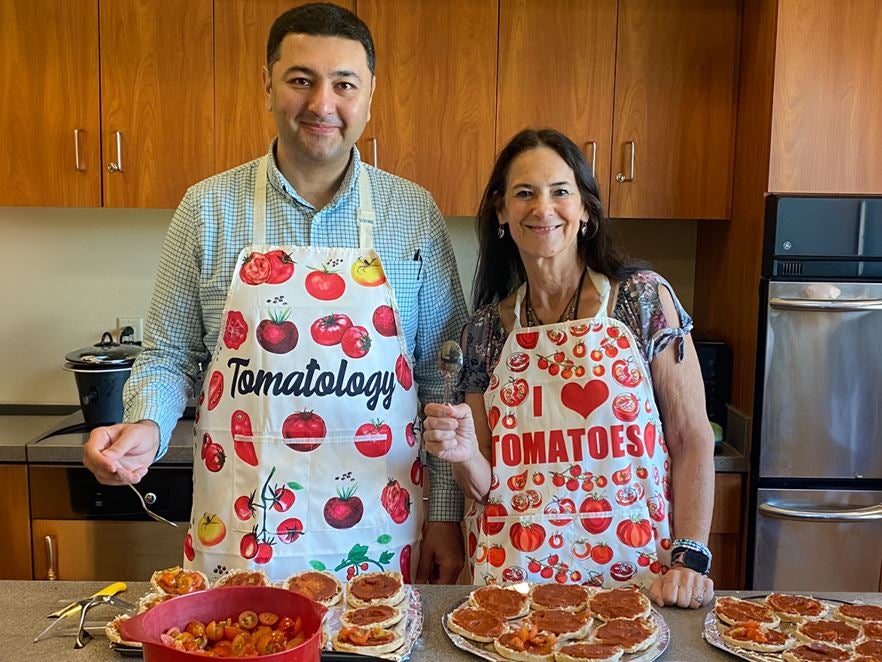 Jenny Ovide, RN, of Community Medicine at Ascension All Saints Family Health Center, stands in a kitchen with a colleague, both wearing tomato-themed aprons and preparing food during a healthy cooking class, with ingredients and prepared dishes on the counter.