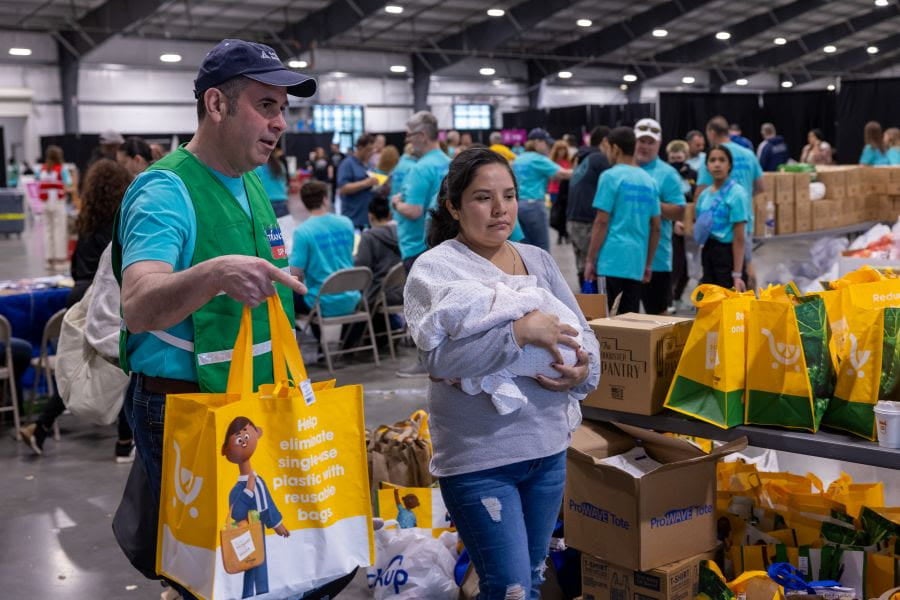 Volunteer in a green vest hands a yellow reusable bag to a woman holding a baby inside a large warehouse filled with people, tables, and boxes of supplies during a community distribution event.